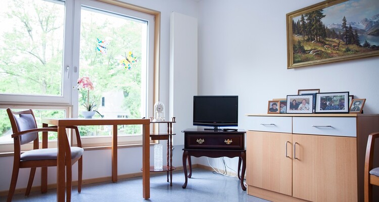 Blick in ein leeres Einzelzimmer im Altenpflegeheim AGAPLESION BETHANIEN LEIPZIG. Vor einem Fenster stehen Tisch und Stuhl, daneben ein Fernseher sowie ein kleines Regal mit Fotos. An der Wand hängt ein Bild.