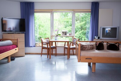Blick in ein leeres Doppelzimmer im Altenpflegeheim AGAPLESION BETHANIEN LEIPZIG. Vor einem Fenster stehen Tisch und Stühle, daneben ein Fernseher auf einem Regal. Im Zimmer stehen außerdem zwei Einzelbetten.